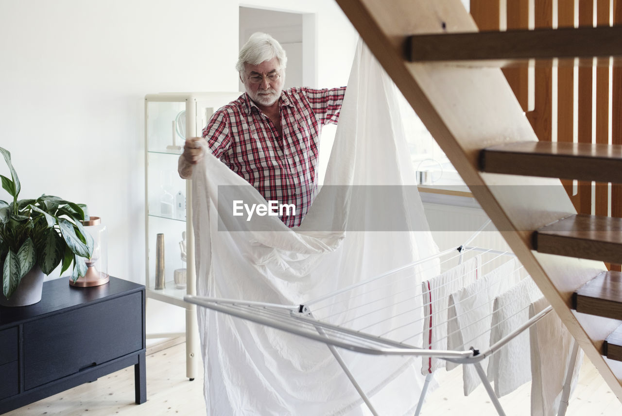 Senior man folding white sheet for drying on rack in living room