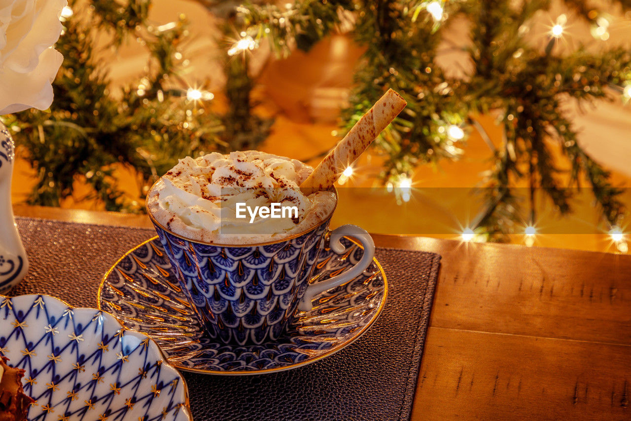 Cup of hot chocolate in a fancy bone china cup with gold and a silver spoon in front of a christmas