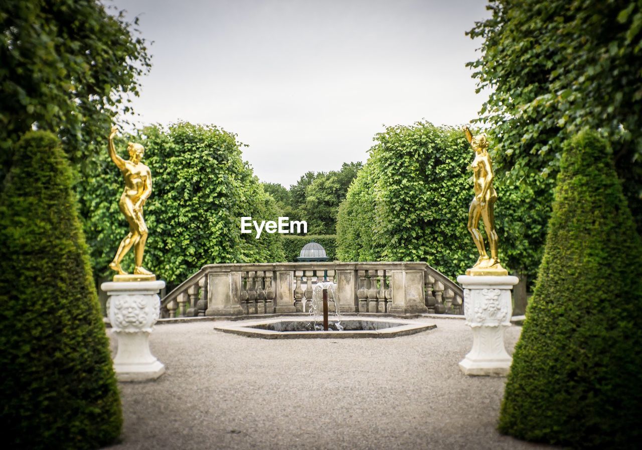 Low angle view of statue and trees with hedges against sky in park