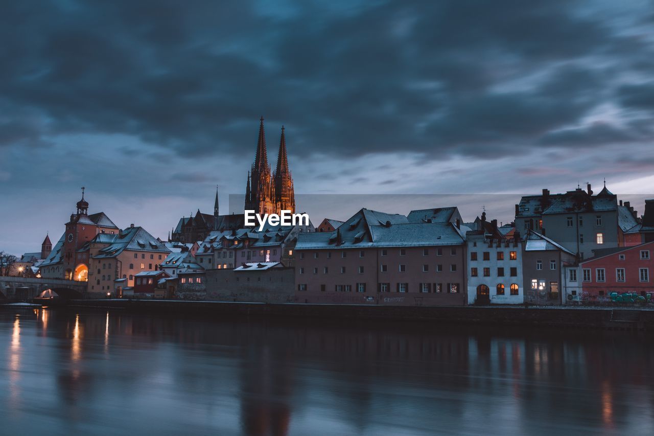 Buildings by river against cloudy sky