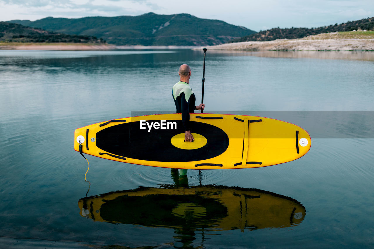 Side view of unrecognizable male surfer in wetsuit standing with yellow sup board and paddle in sea water
