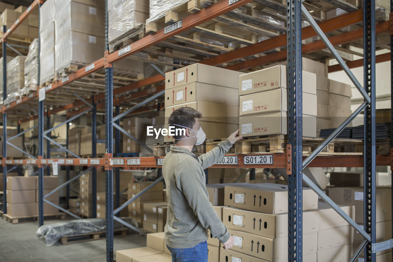 Man wearing mask holding boxes on shelf in warehouse