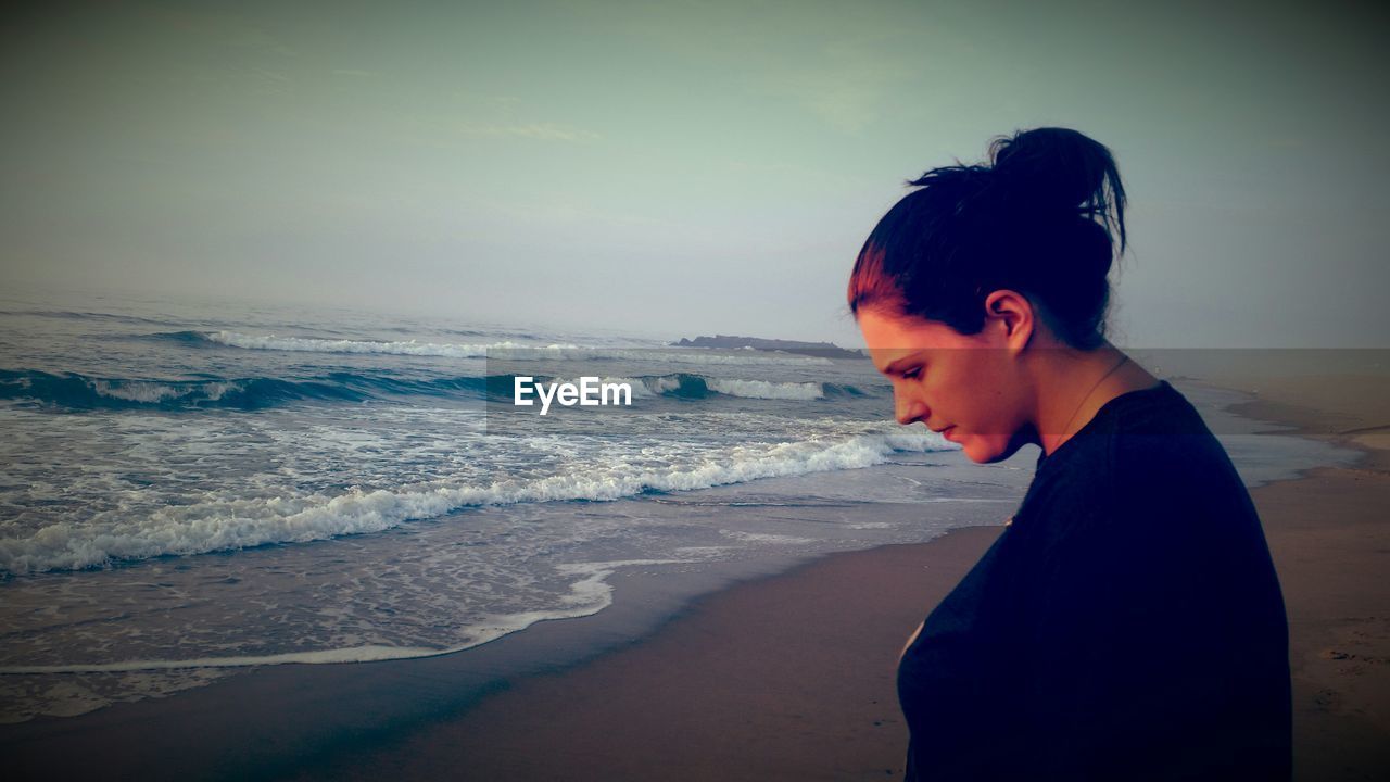 Woman looking down while standing at beach against sky