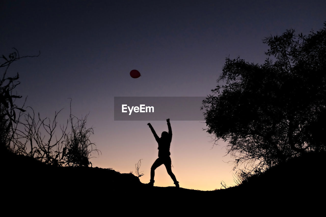 Silhouette of woman trying to catch balloon against sky during sunset