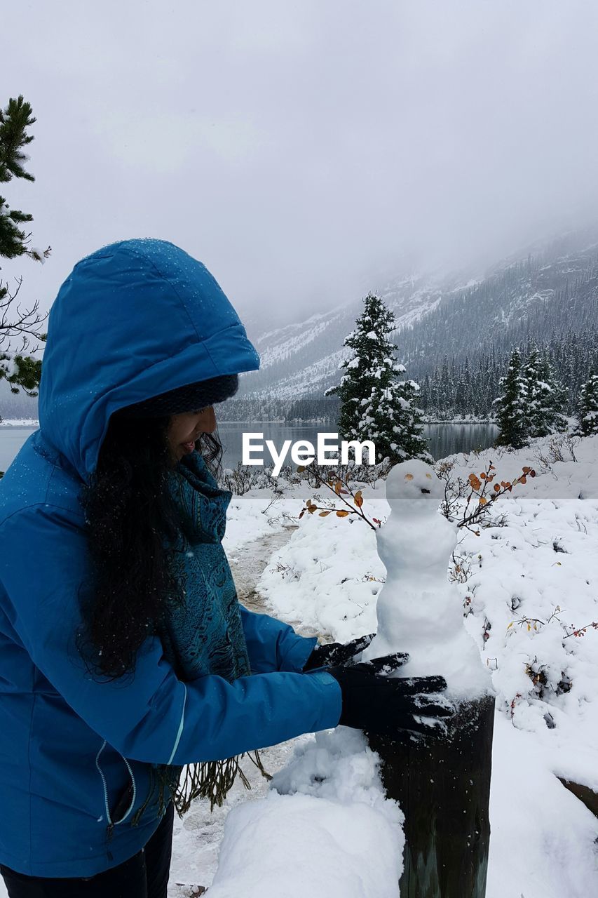 Woman making snowman on wooden post against lake and mountains