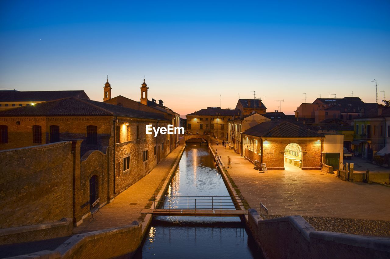 Illuminated canal amidst buildings against clear sky at sunset