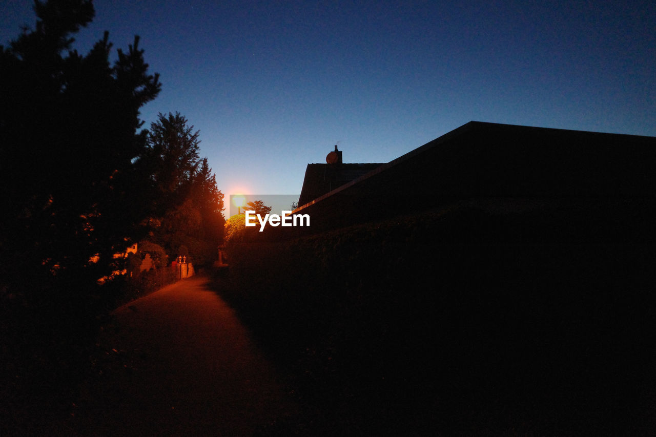 Silhouette of suburban buildings against clear sky at sunset with streetlamp illuminating a pathway 