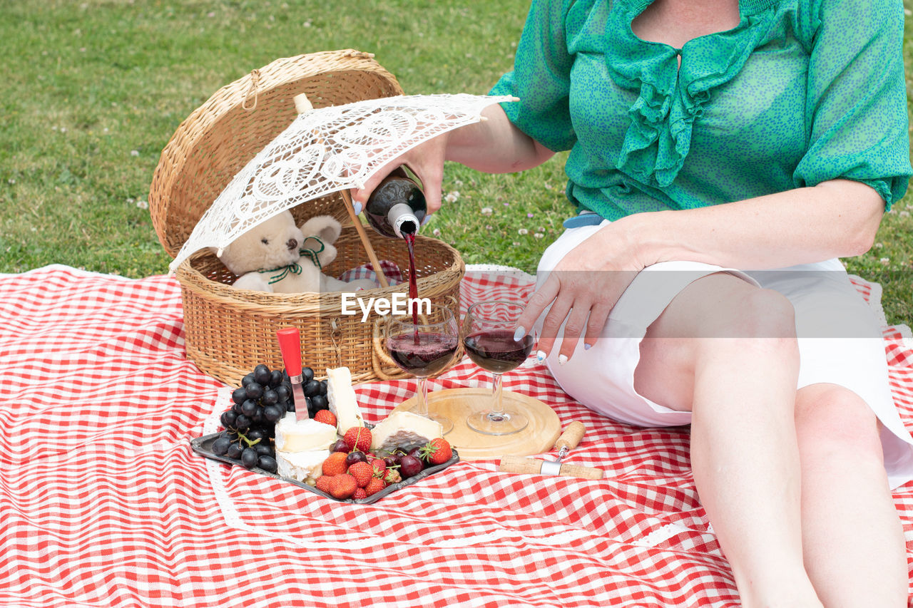 Woman in a green blouse sits on a red checkered picnic rug, red wine and chees