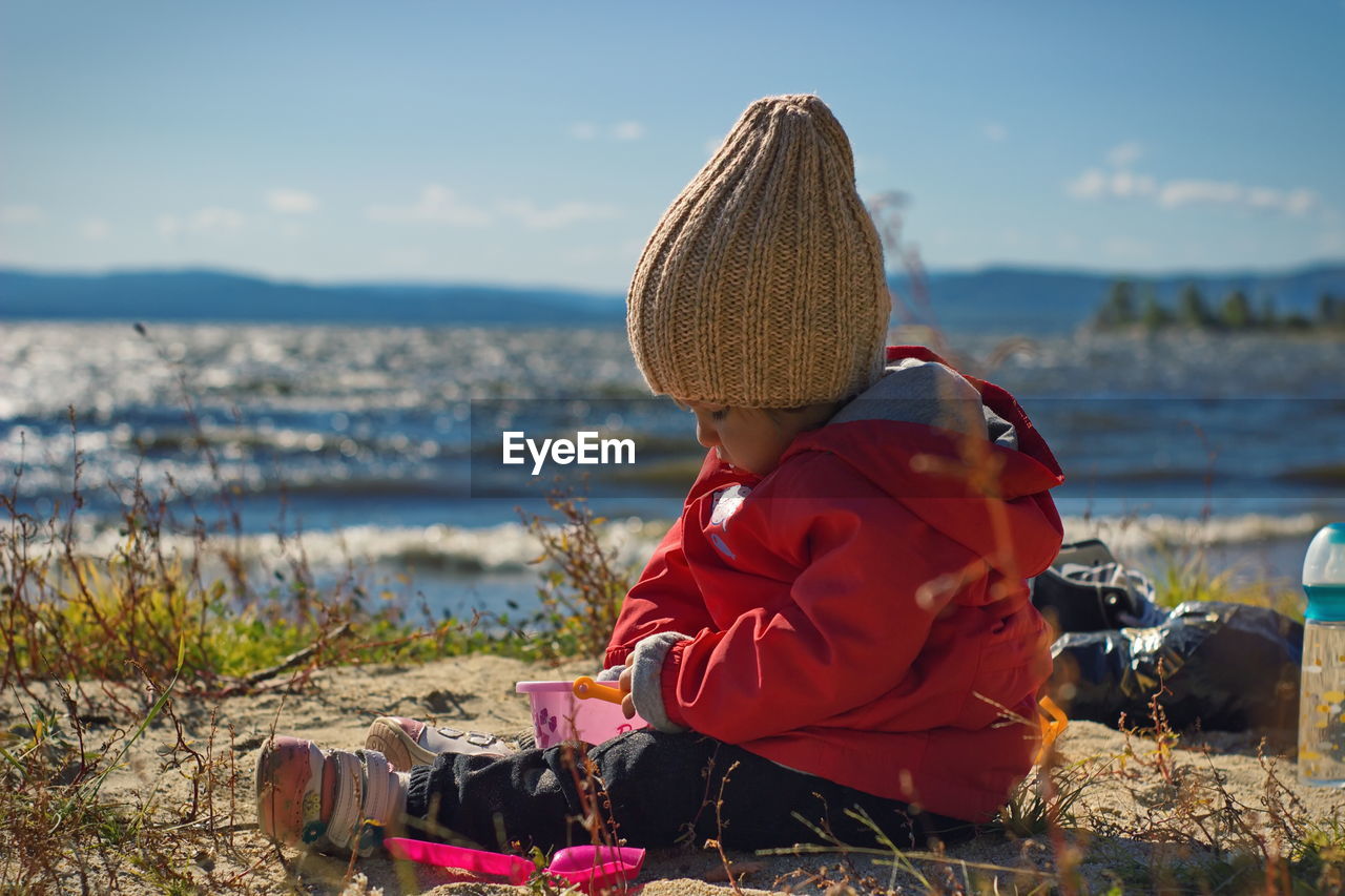 Side view of baby girl playing with sand while sitting on beach against blue sky