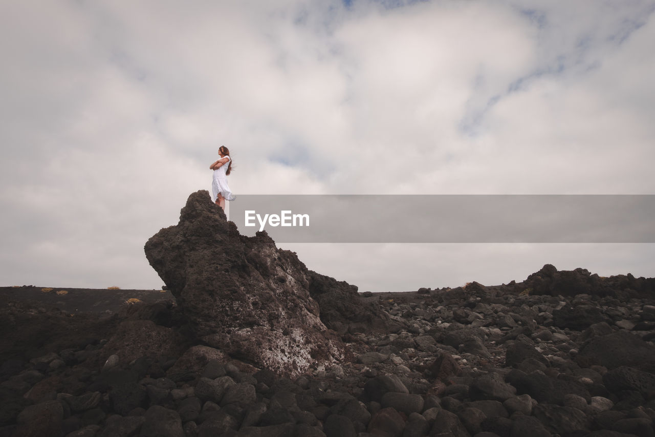 Low angle view of young man standing on rock against cloudy sky