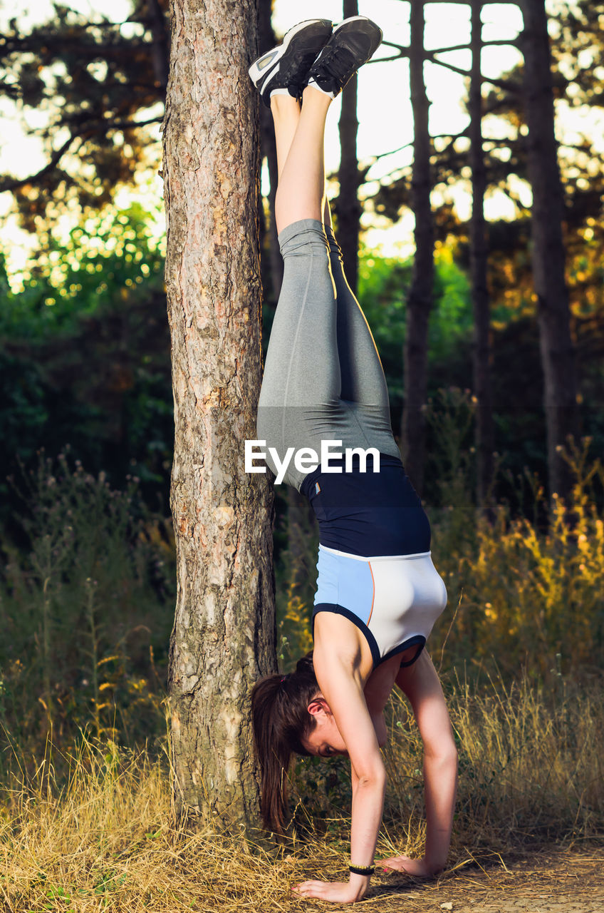 Young woman doing handstand on field against trees in forest during sunset