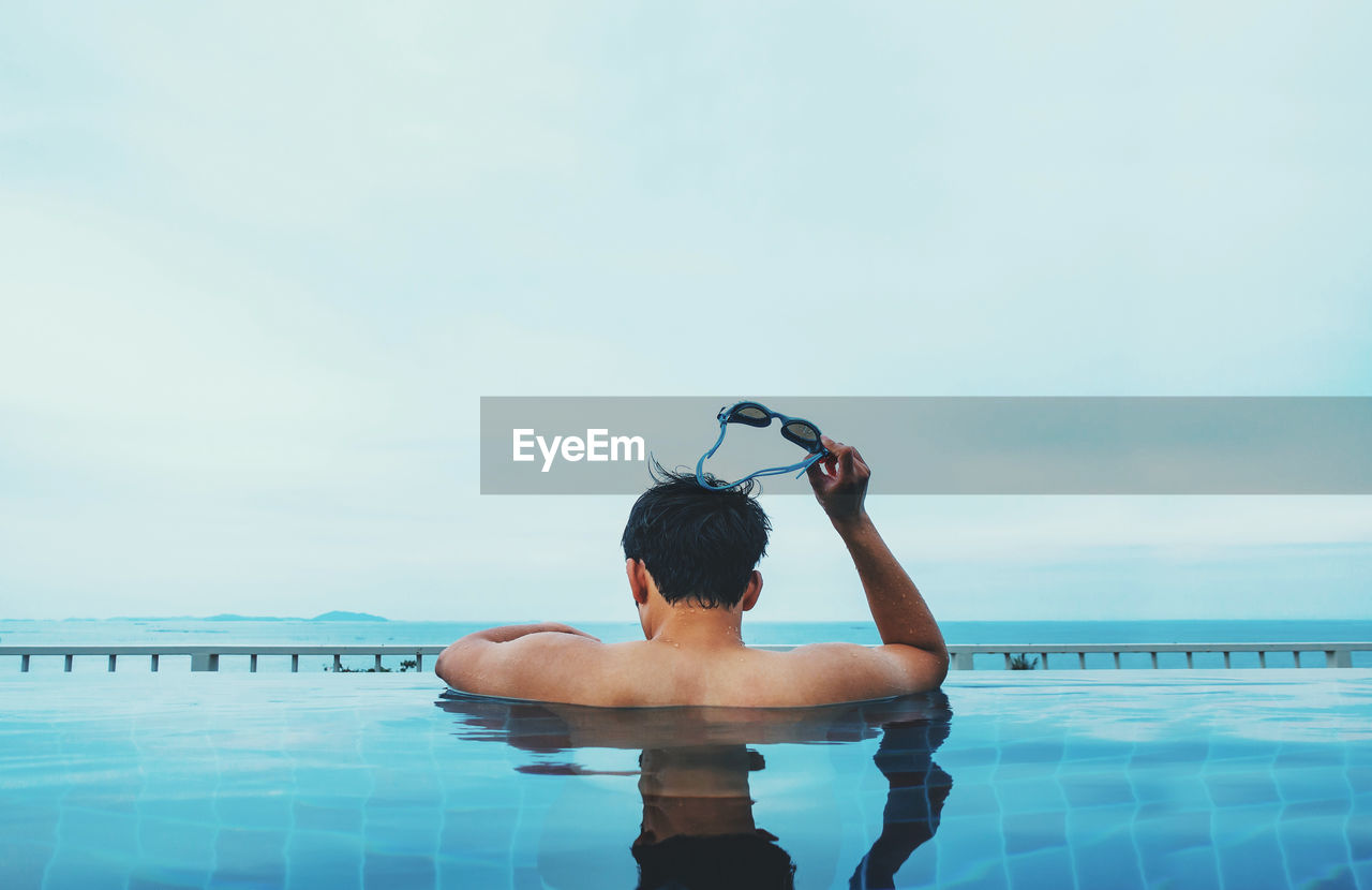 Rear view of shirtless man holding swimming goggles in swimming pool against sky