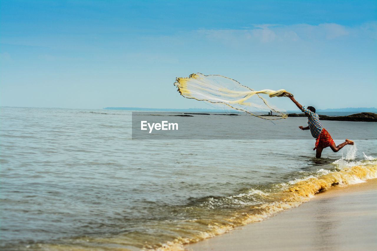 Full length of man throwing fishing net in sea against blue sky