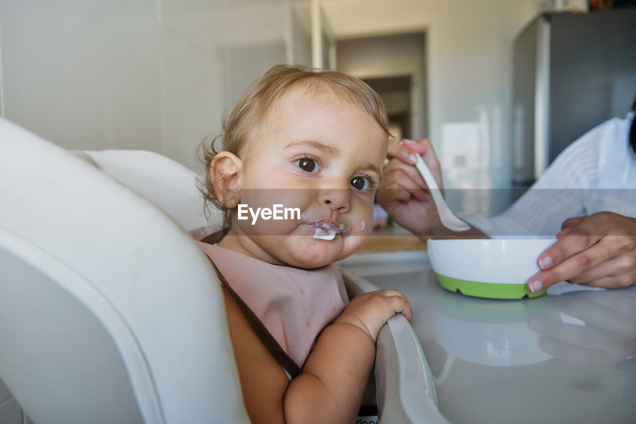 Little girl sitting on a high chair with a bib eating from the spoon with cropped unrecognizable mother feeding her