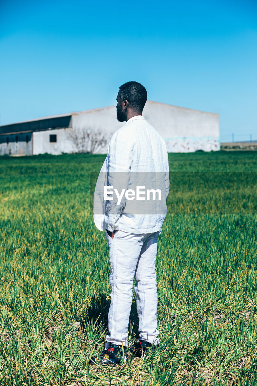 Back view of black male model in trendy white suit and authentic african shirt with frown while standing in grassy field