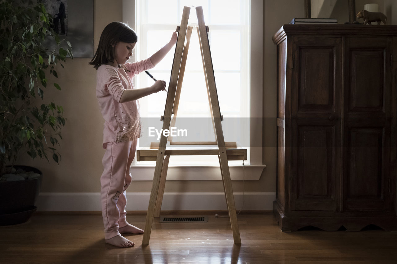 Side view of girl drawing on artist's canvas while standing at home