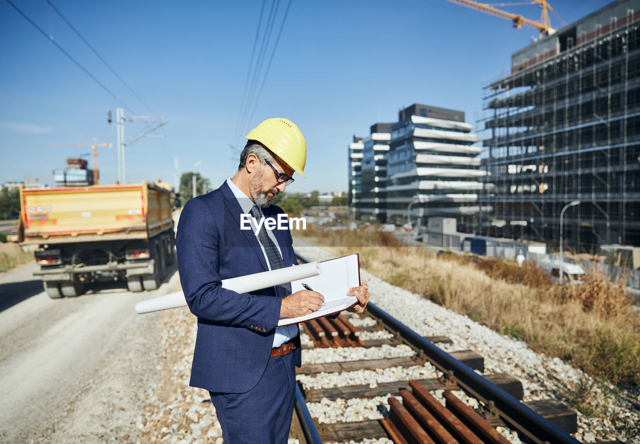 Side view of engineer writing on book while standing against blueprint