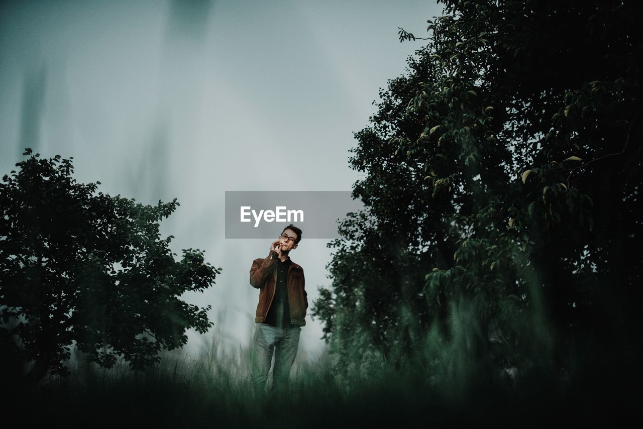 Portrait of young man smoking while standing by trees against sky