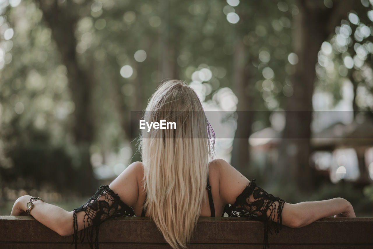 Rear view of woman sitting on bench with arms outstretched at park