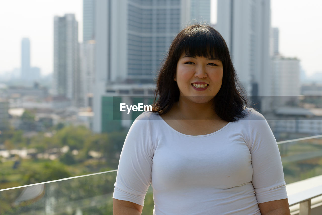 PORTRAIT OF SMILING YOUNG WOMAN AGAINST CITYSCAPE