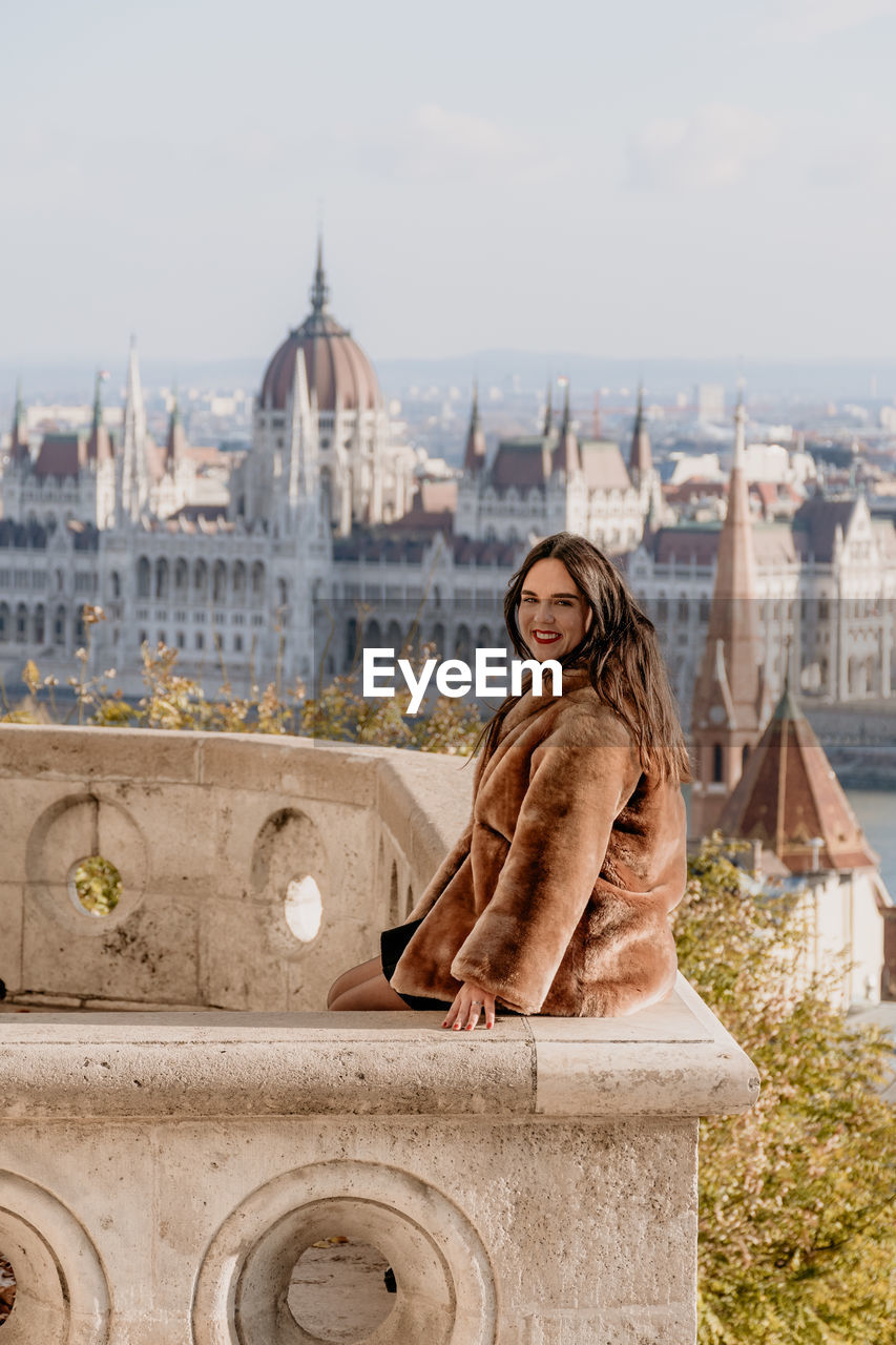 Portrait of beautiful young woman on balcony overlooking hungarian parliament in budapest, hungary