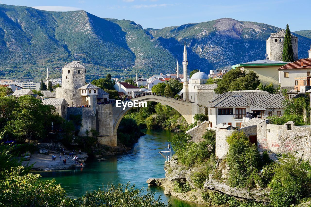 Bridge over river by houses and mountains against sky