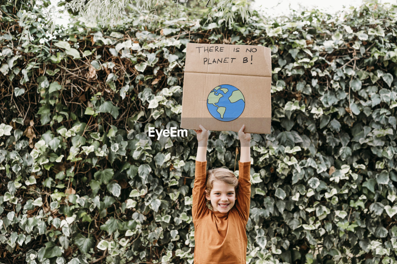 Happy girl with arms raised holding banner with message and planet drawing in front of plant