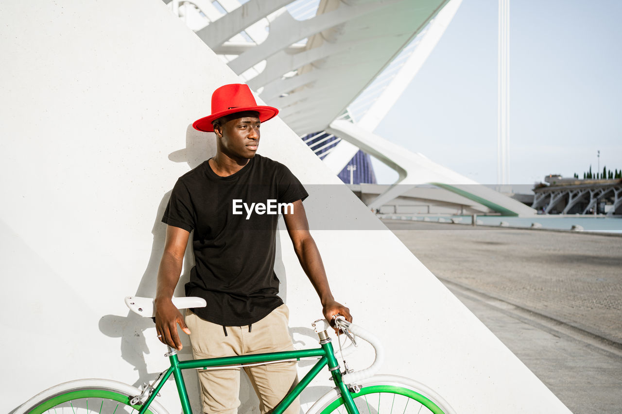 Confident african american male in trendy clothes standing with modern bike in urban area and looking away