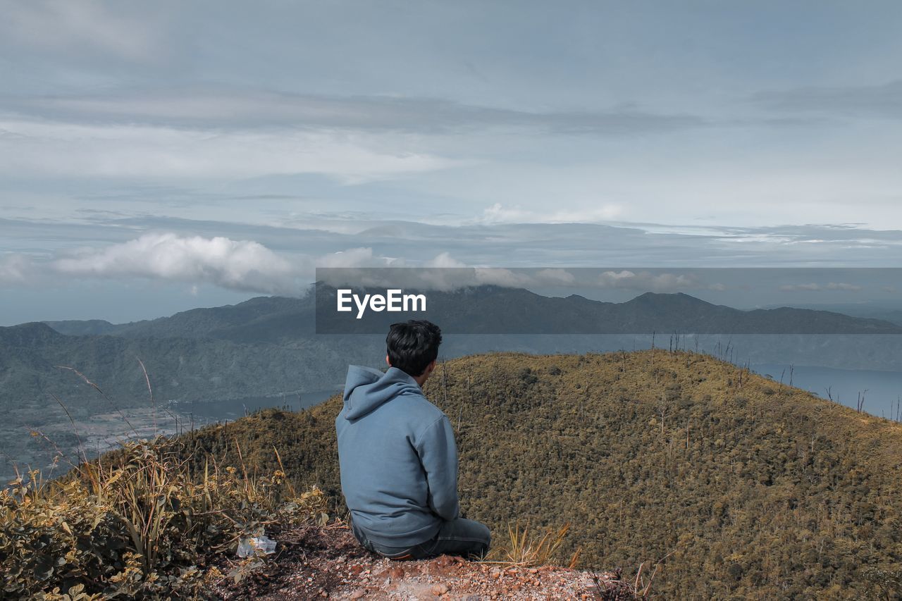 Rear view of man sitting on mountain against sky.