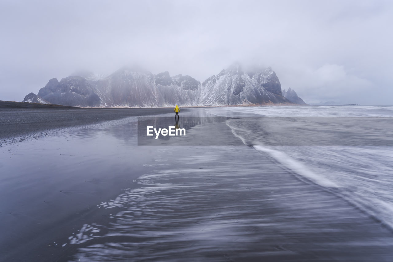 Distant view of unrecognizable traveler standing on wet black sand beach near waving sea against mountain ridge and cloudy sky in iceland