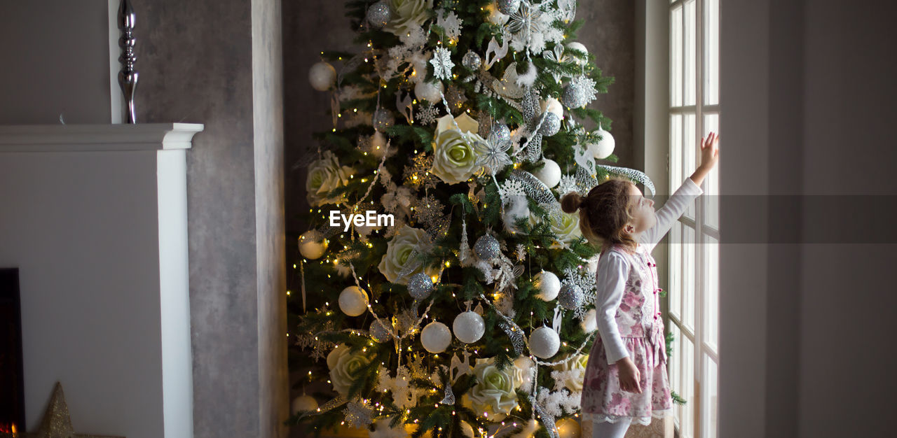 WOMAN STANDING BY CHRISTMAS TREE IN WINDOW