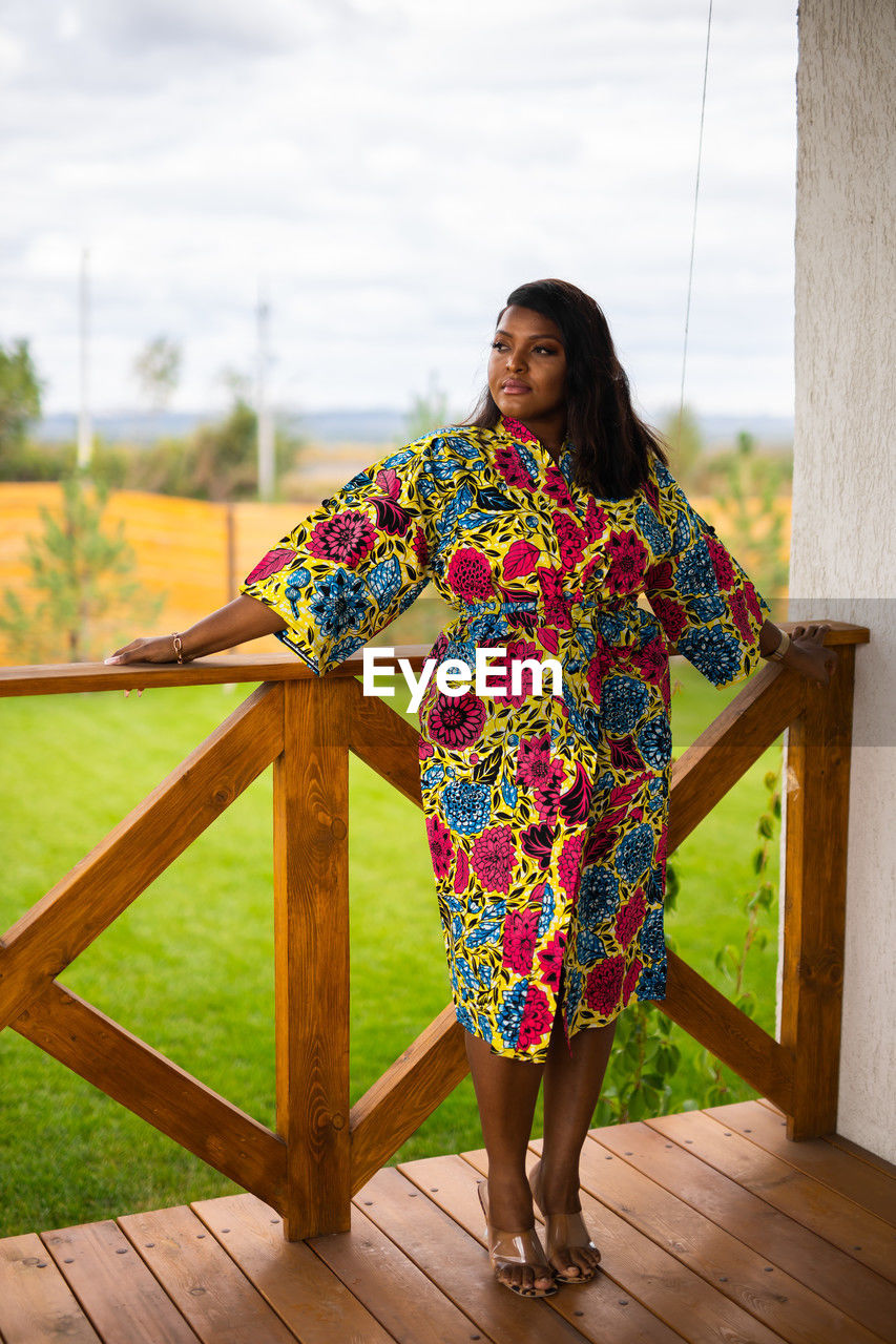 one person, spring, women, adult, dress, full length, clothing, standing, yellow, young adult, smiling, nature, wood, fashion, lifestyles, pattern, sky, architecture, leisure activity, portrait, happiness, female, hairstyle, floral pattern, outdoors, day, front view, casual clothing, emotion, relaxation, looking, brown hair, flower, multi colored, cloud, photo shoot, long hair, looking at camera, plant, cheerful