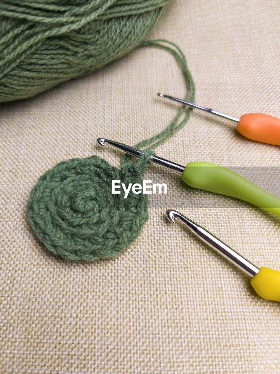 Green yarn thread with and three colorful hooks for knitting and hobbies on gray fabric background.