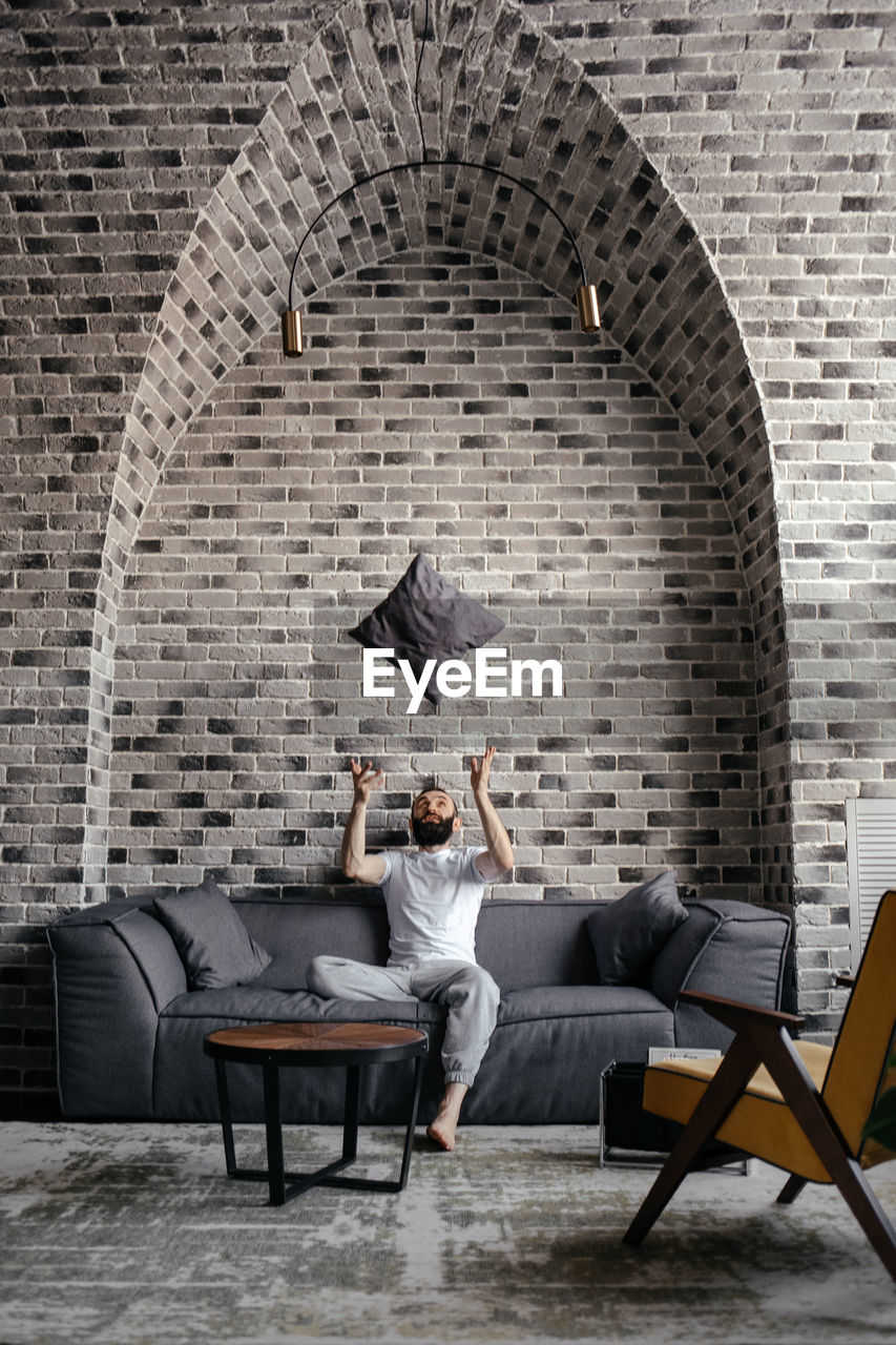 A man with a beard sitting on the sofa and throwing up a pillow