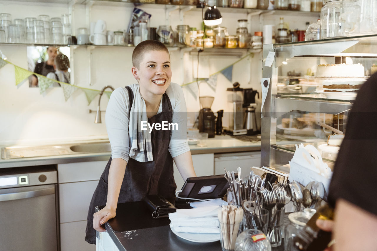 Happy female owner leaning on counter while looking at colleague in cafe