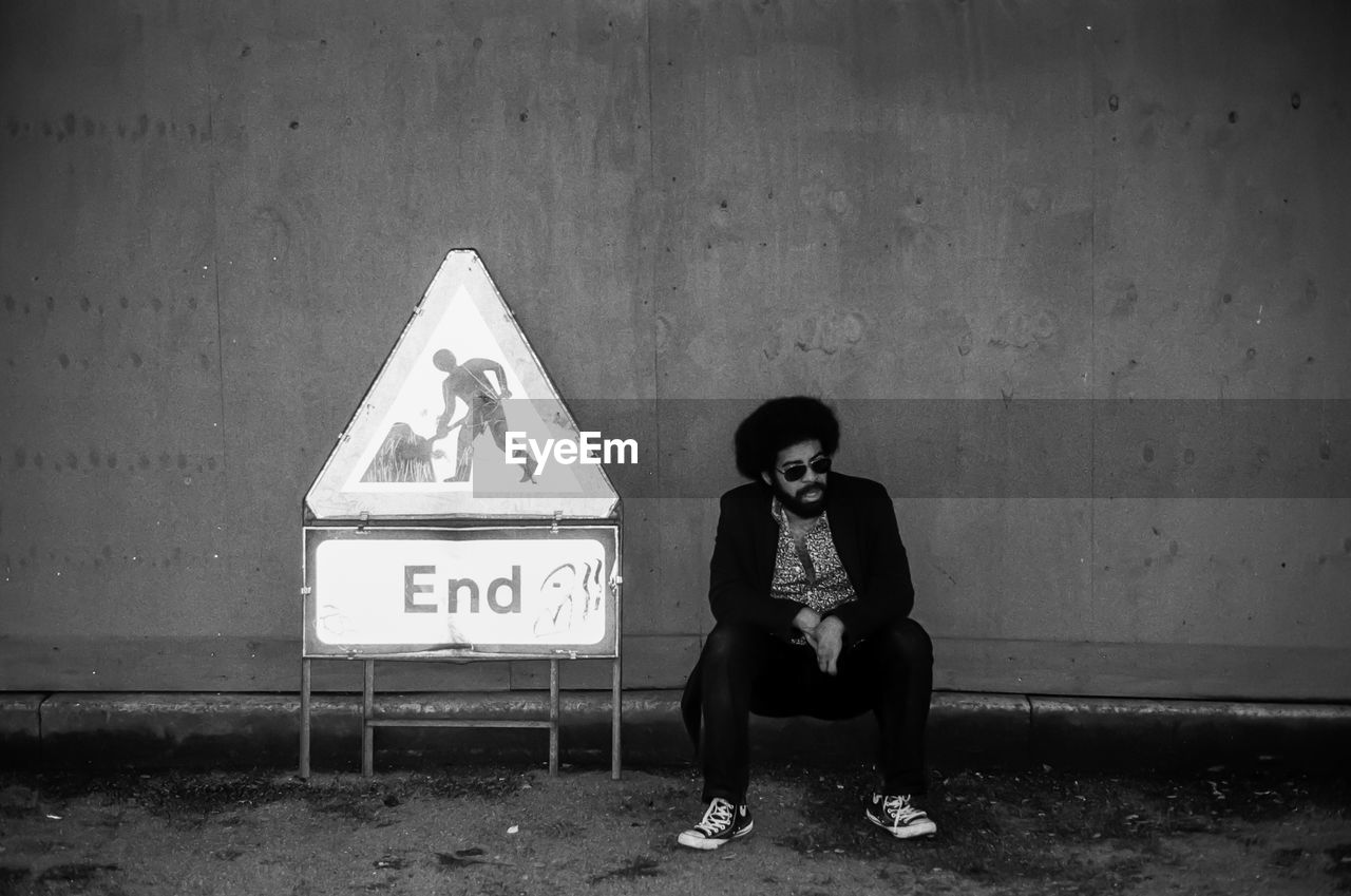 Bearded man sitting by sign against wall