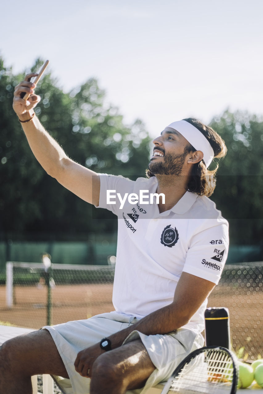 Smiling man taking selfie through smart phone while sitting on bench at tennis court