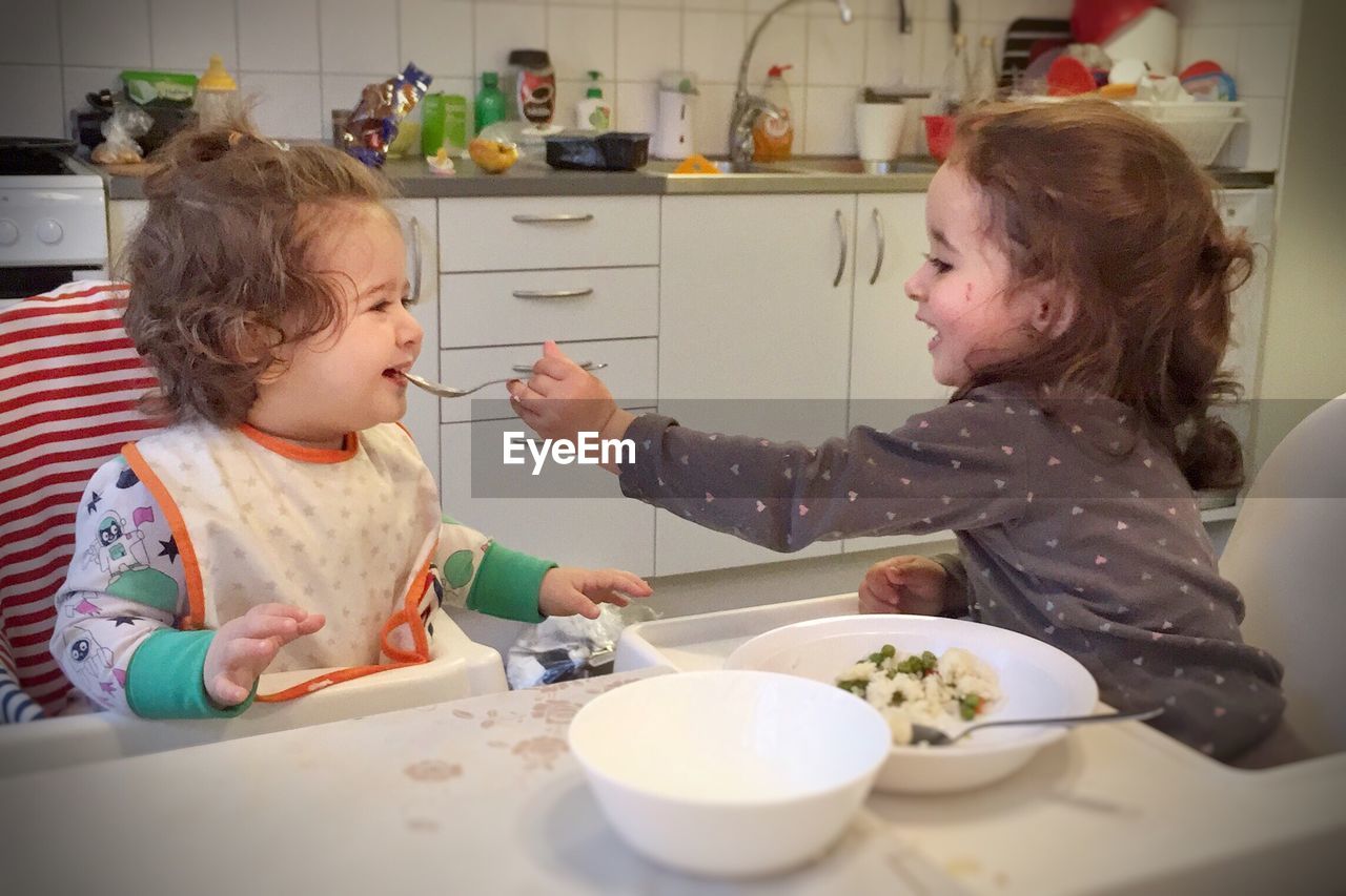 Side view of girl feeding sister while sitting on high chair