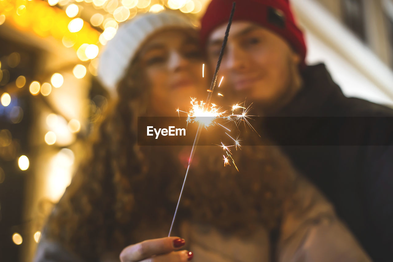 A sparkler burns in the hands of a girl hugged by her boyfriend in a christmas mood.