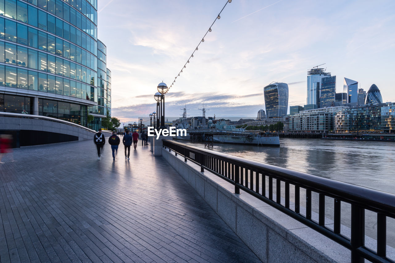 People walking by more london riverside buildings, and walkie-talkie tower in twilight.