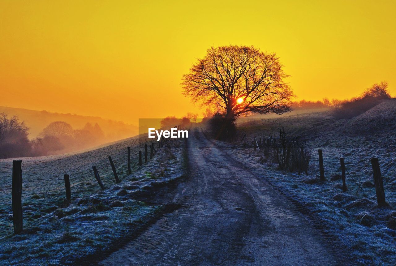 DIRT ROAD AMIDST SNOW COVERED FIELD AGAINST SKY DURING SUNSET