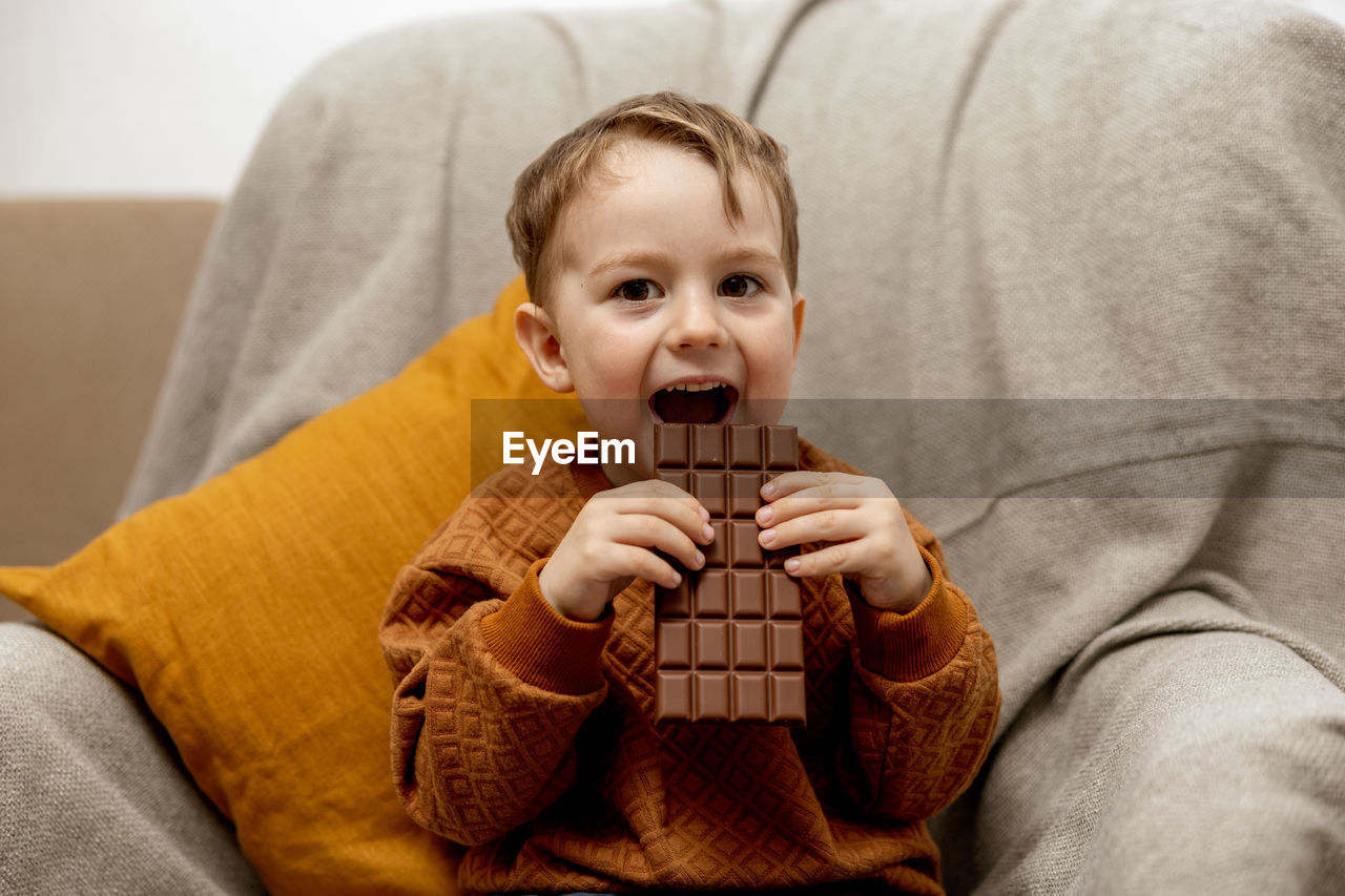 Little adorable boy sitting on the couch at home and eating chocolate bar. child and sweets