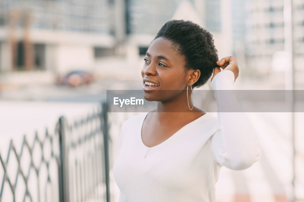 PORTRAIT OF SMILING YOUNG WOMAN STANDING AGAINST CITY