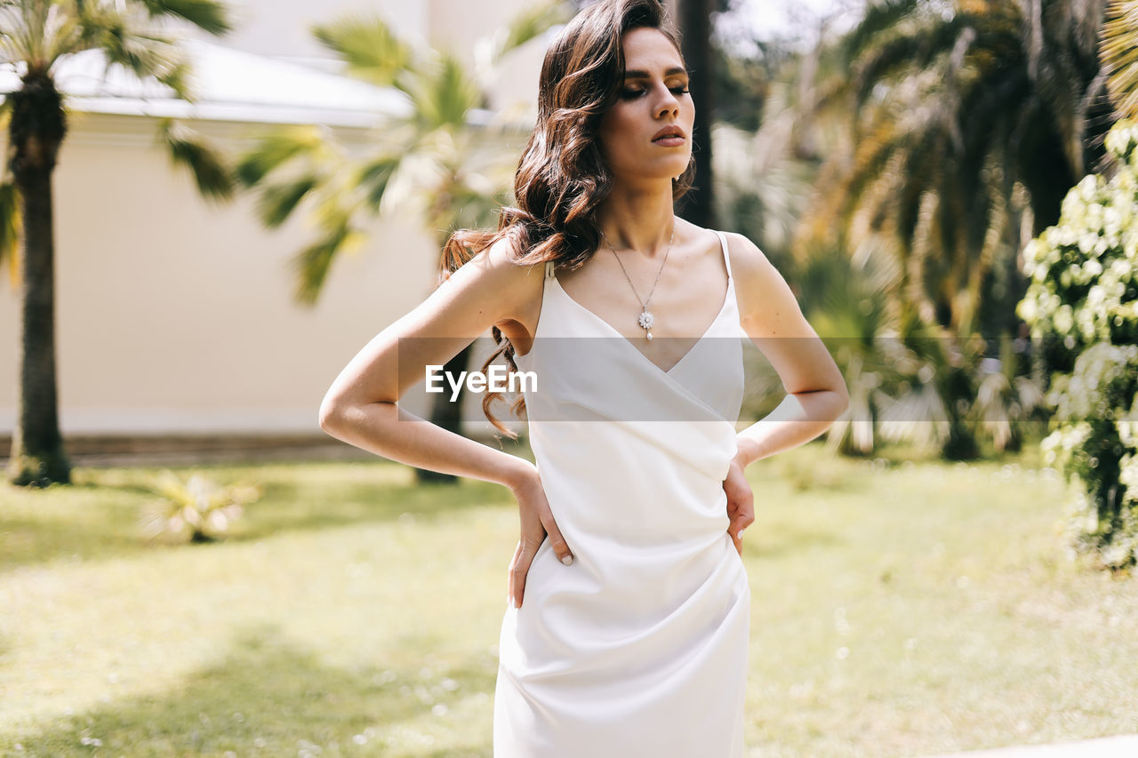 An elegant young woman bride in a wedding dress walks through a green park among plants and trees