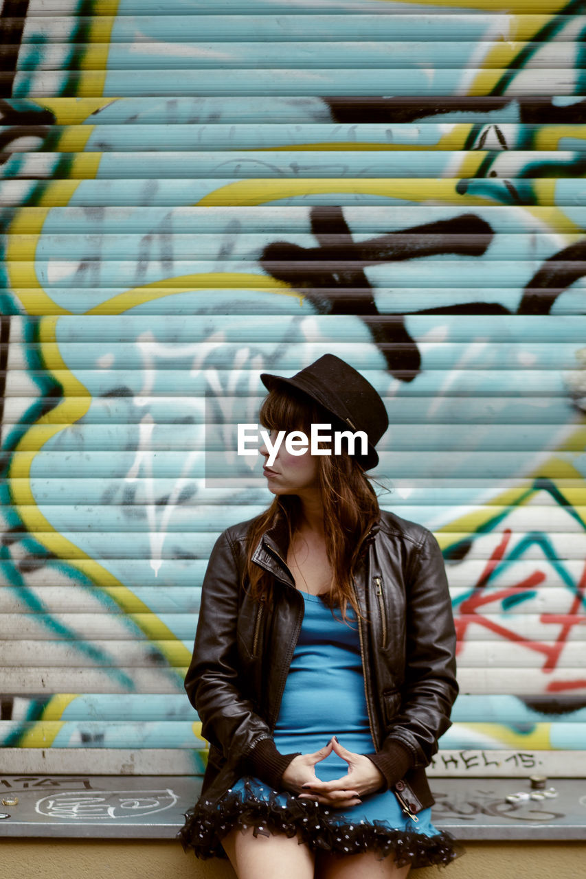 Woman wearing hat looking away against graffiti shutter in city