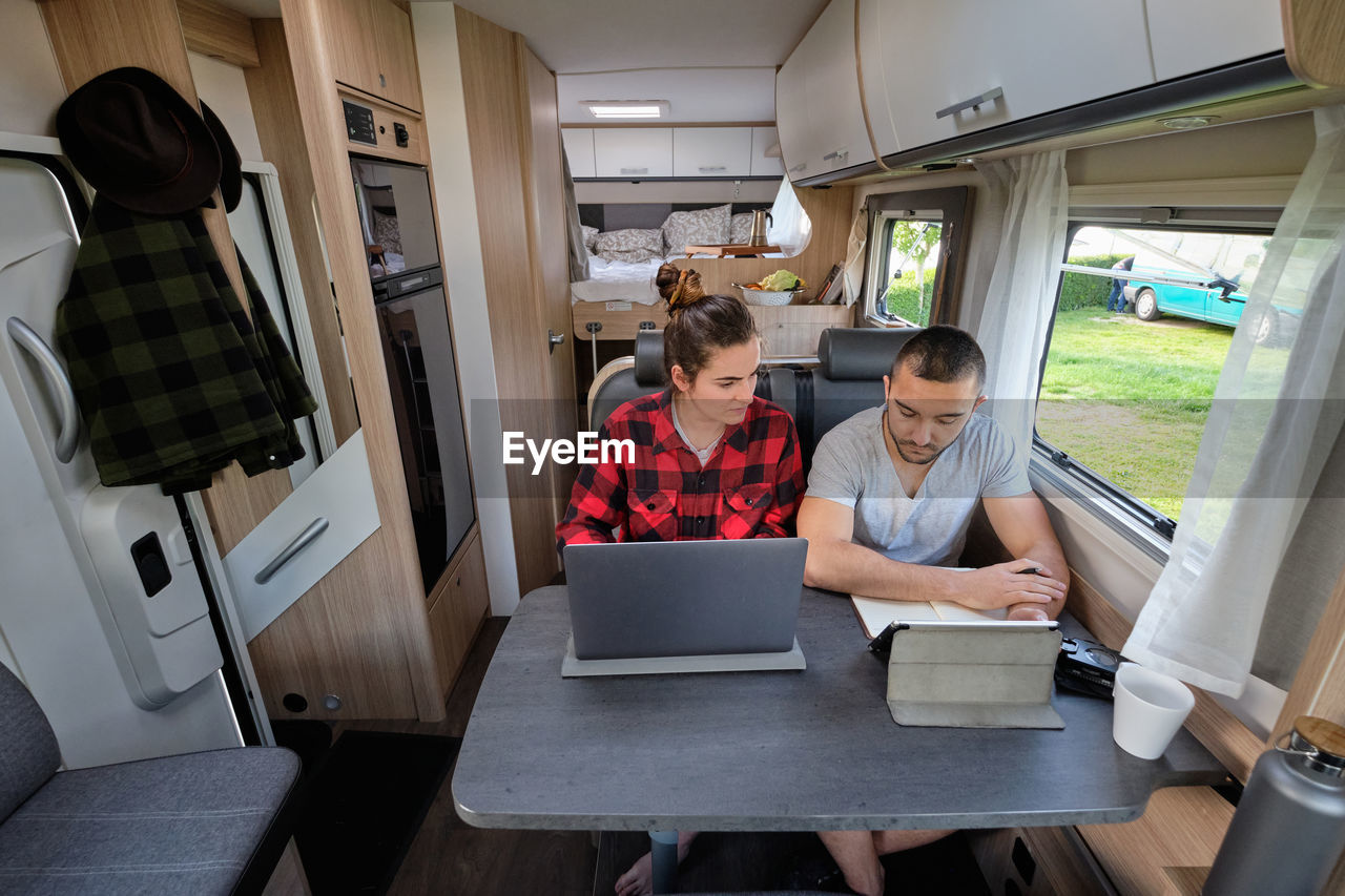 Smiling traveling couple of freelancers using laptop and tablet while working on project remotely at table in caravan