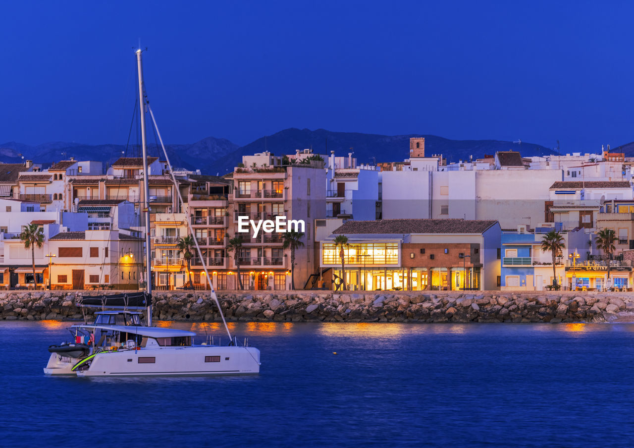 Sailboats in sea by buildings against blue sky