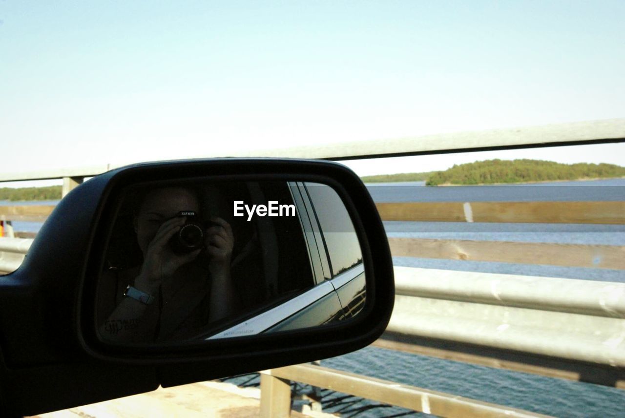 Reflection of woman with camera on side-view mirror of car on bridge