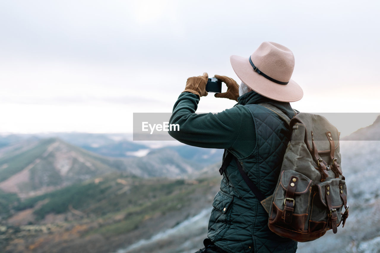 Side view of unrecognizable senior male explorer taking photo of mountainous landscape while using smartphone and enjoying vacation in caceres
