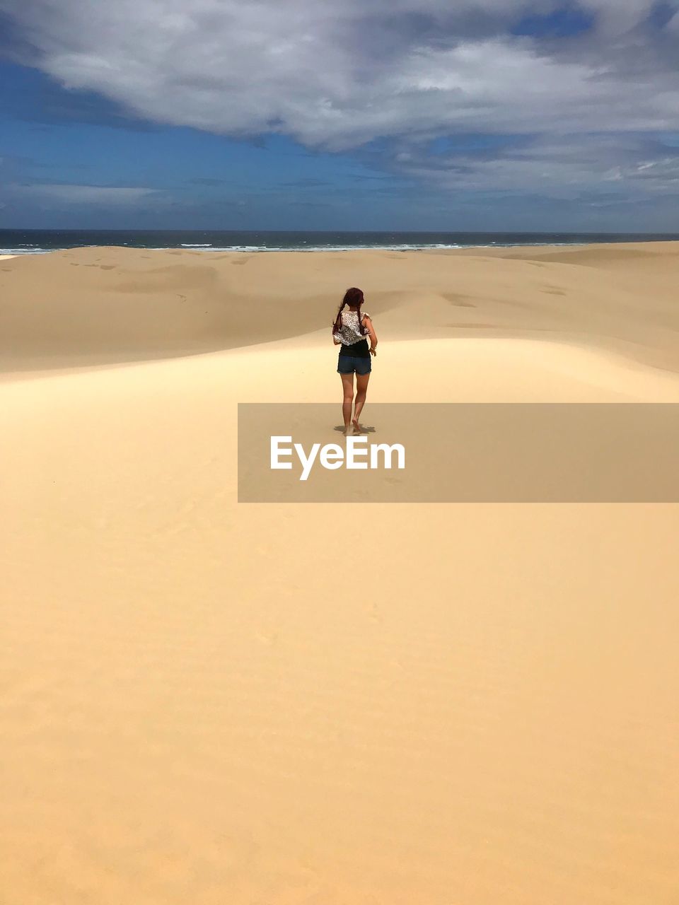 Rear view of woman running on sand dune at beach against cloudy sky